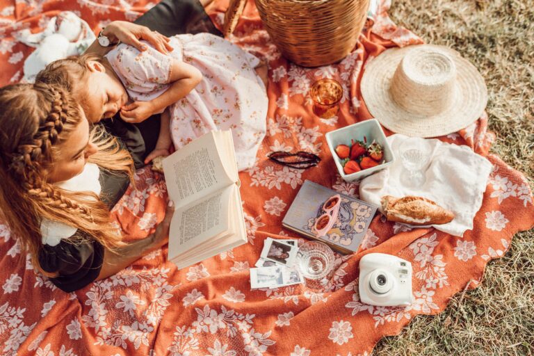 A mother and daugher sat on an orange picnic blanket reading a book whilst cuddled upto each other