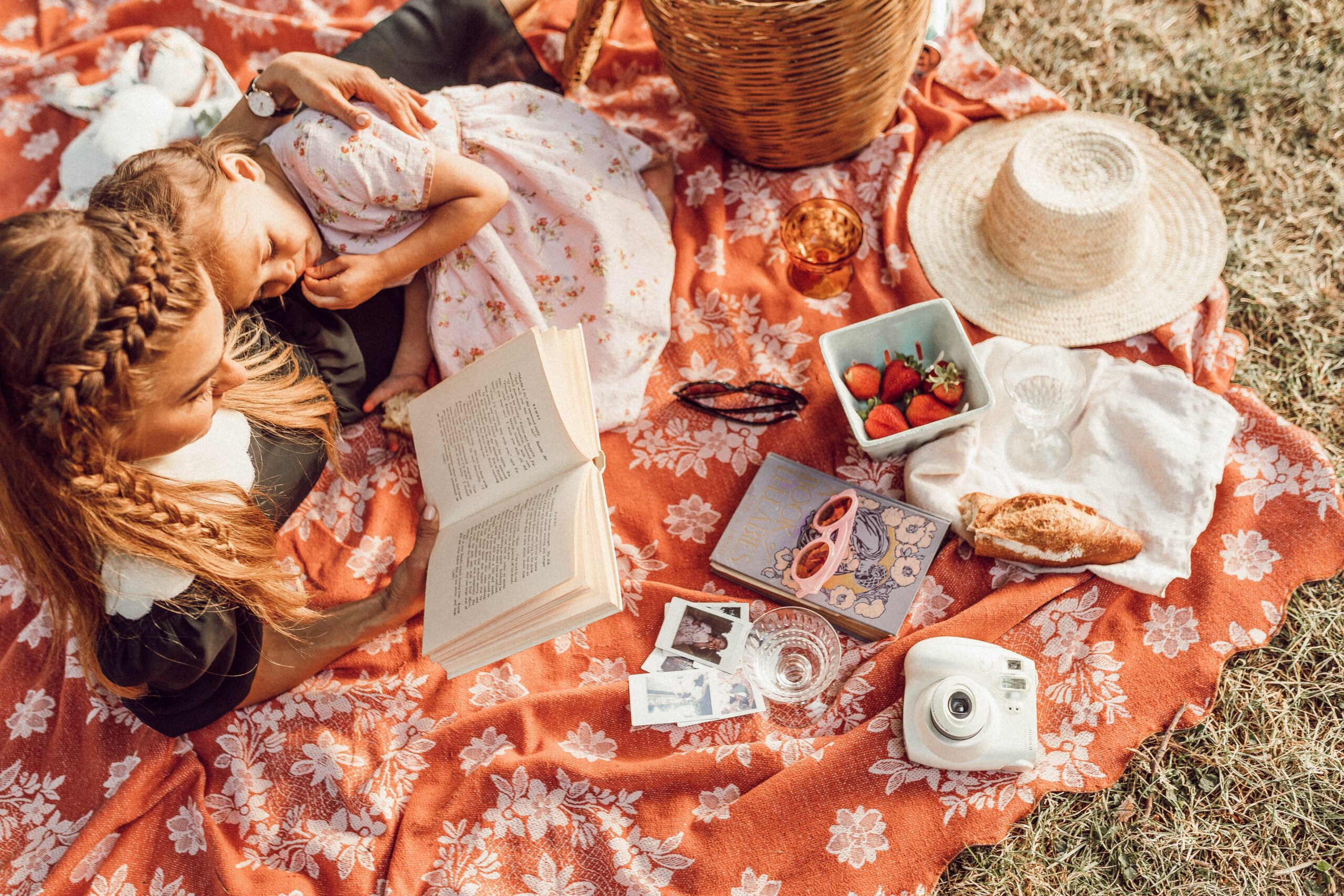 A mother and daugher sat on an orange picnic blanket reading a book whilst cuddled upto each other