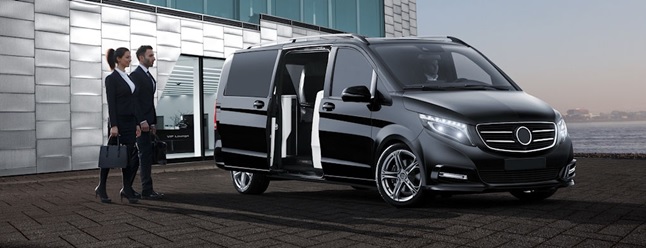 Two people in business attire walk towards a black luxury van with sliding doors open, parked on a waterfront promenade beside a modern glass building in Madrid.