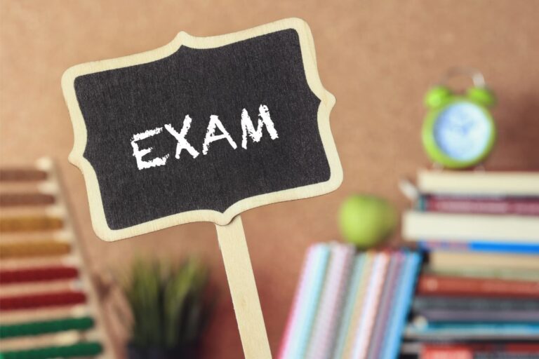 A small blackboard with the word "EXAM" written on it is held up in front of a blurred background featuring books, an abacus, a green apple, and an alarm clock—symbols of preparation for forthcoming exams.