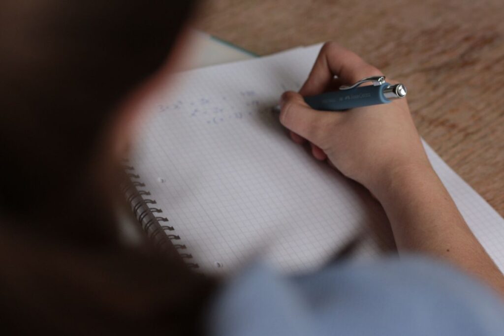 A person writes with a blue pen on a spiral-bound notebook with squared paper, working on mathematical problems—perhaps preparing for upcoming exams. The focus is on the hand and paper, whilst the background is blurred.