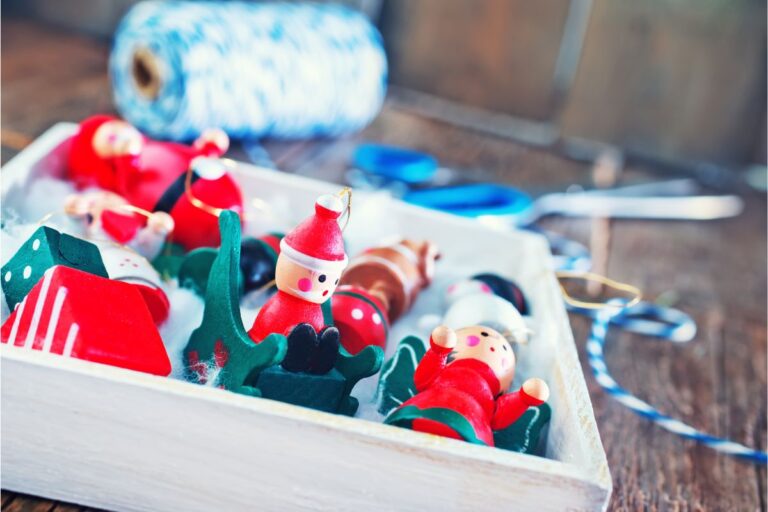 A white tray holds small wooden Christmas decorations—perfect things to buy before Christmas—including Father Christmas figures and green trees, with a reel of blue and white string and scissors blurred in the background on a wooden table.