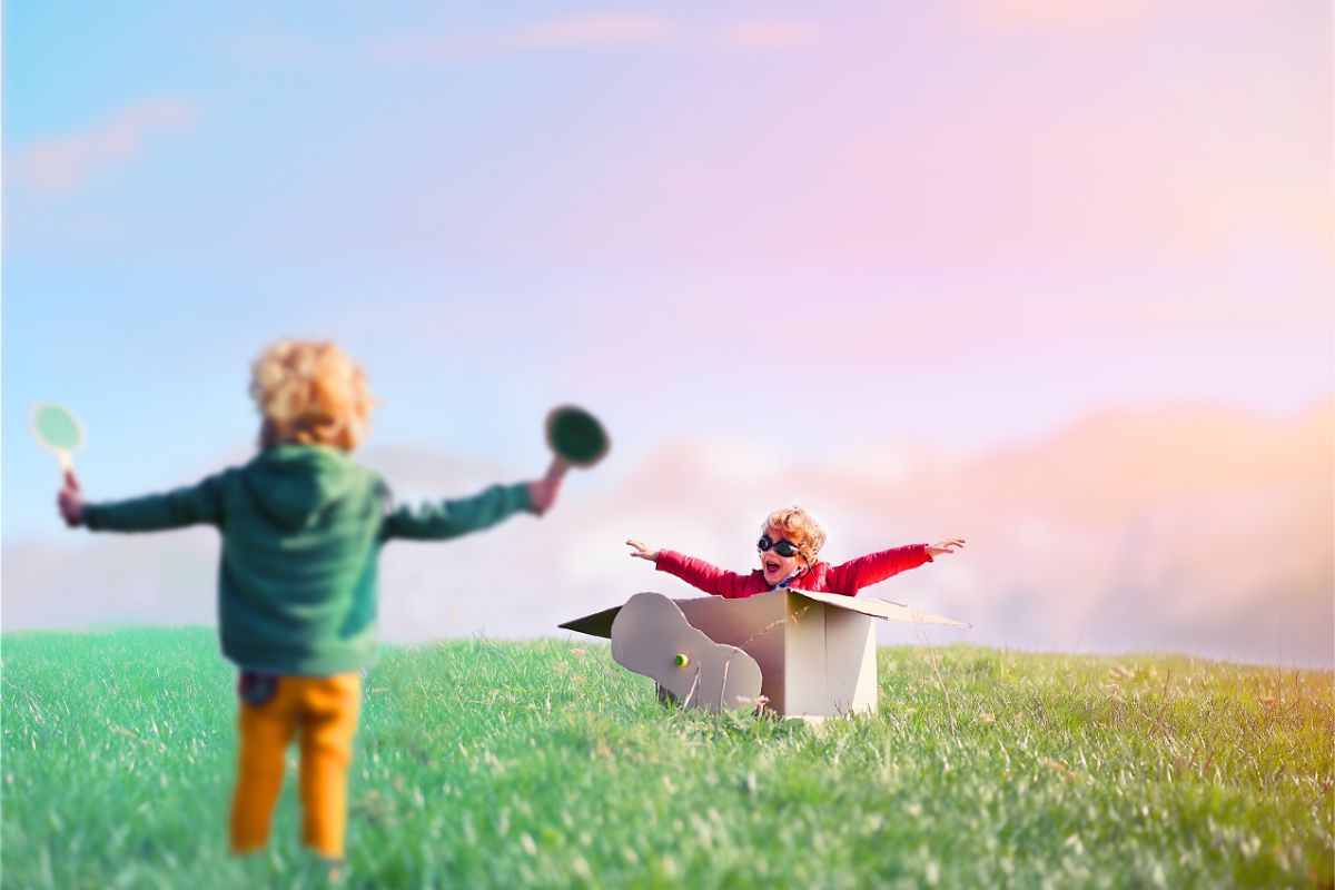 Two children play outside on a grassy field; fuelled by imagination, one stands with arms raised holding paddles, while the other, wearing goggles and arms outstretched, pretends to fly in a cardboard box aeroplane under a colourful sky.