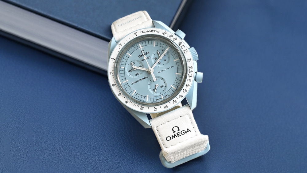 A light blue Omega Speedmaster wristwatch with a white strap rests on a blue surface next to a closed book. Renowned among watches, it features multiple dials and tachymeter markings on its bezel.