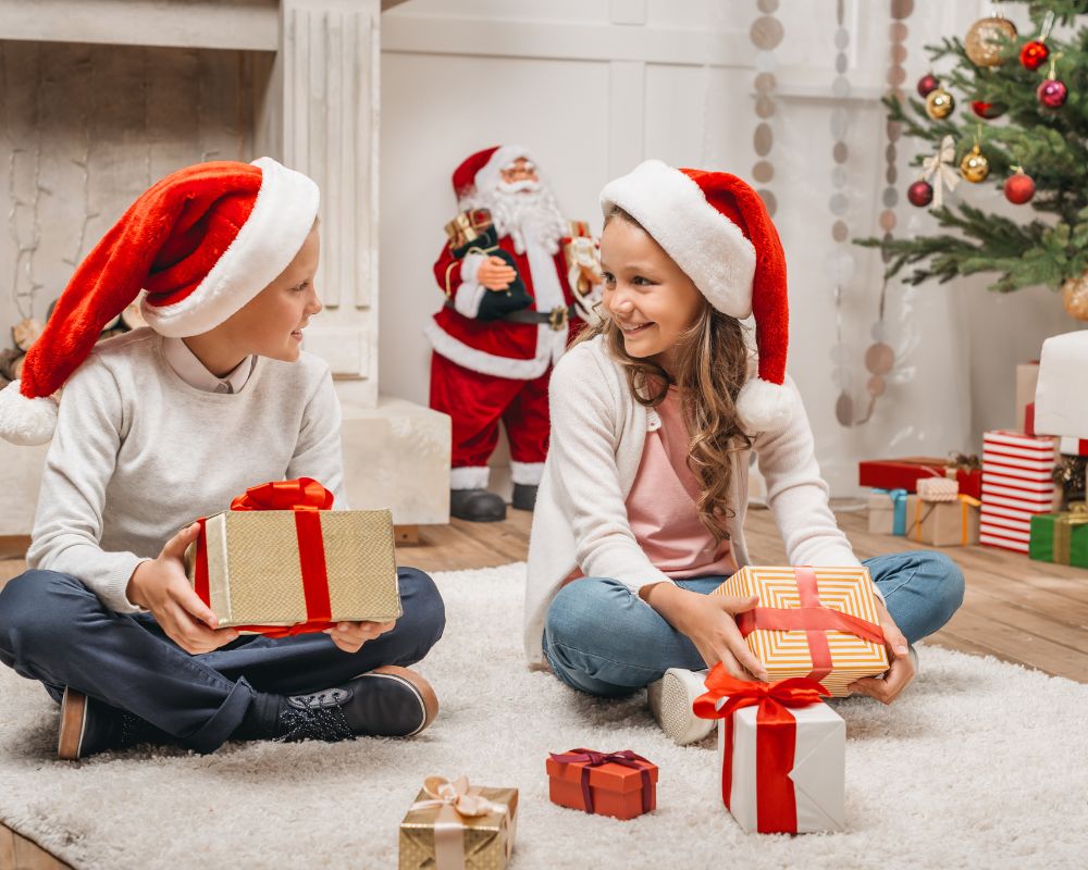 Two children wearing Father Christmas hats sit on the floor by a Christmas tree, holding presents and smiling at each other. A Father Christmas decoration and more presents are in the background.