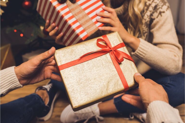 Two adults sit on the floor exchanging wrapped presents; one holds a gold box with a red ribbon, the other has a red-and-white striped present. Both are dressed in casual clothes and cosy jumpers.