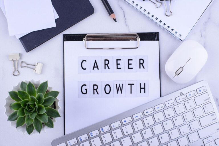 A clipboard with the words "Career Growth" on paper tiles, surrounded by a keyboard, computer mouse, potted succulent, pencil, paper clips, and notebooks—everything you need for planning your career on a white desk.