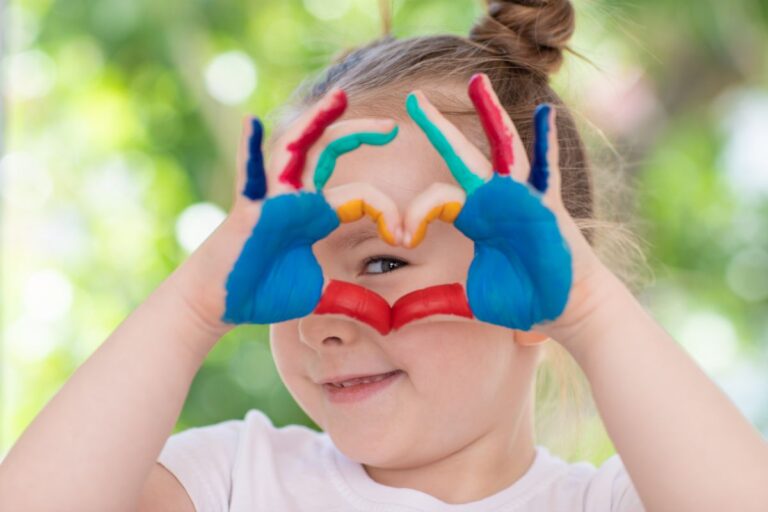 A smiling child showcases their creativity, hands covered in colourful paint, forming a heart shape with their fingers and looking through it. The bright, blurred background highlights the joy of a child's creativity against lush green foliage.