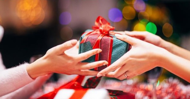 Two people exchange a wrapped present with red ribbon, surrounded by festive lights and blurred holiday decorations in the background.