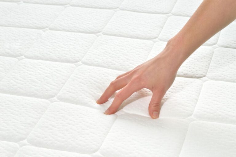 A hand presses down gently on a white, textured mattress surface, showcasing the cushioned mattress sections arranged in neat squares.