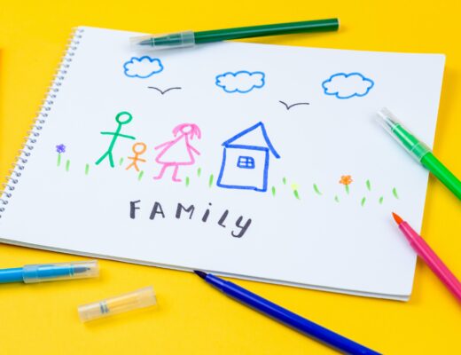 A child’s drawing on white paper shows a stick-figure family of three, a house, grass, flowers, clouds, and birds. The word "FAMILY" is written below. Colourful felt-tip pens are scattered on a yellow background, reflecting joyful family lifestyle moments.