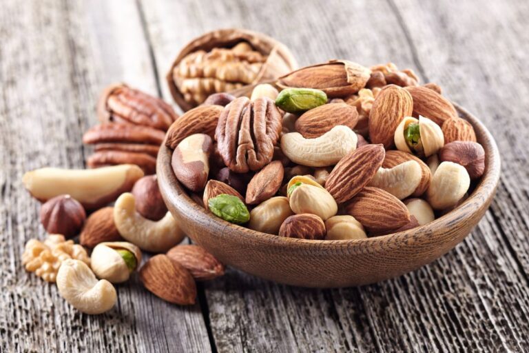 A wooden bowl brimming with mixed nuts—including almonds, cashews, pistachios, walnuts, pecans, and hazelnuts—rests on a rustic wooden surface, showcasing the perfect snack for any nut lover.