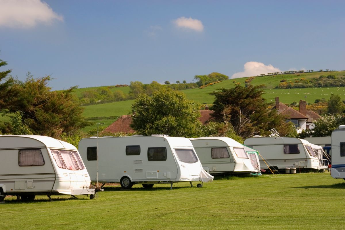Several caravans are parked on a grassy field under a blue sky, perfect for relaxing caravan holidays. Rolling green hills and scattered trees surround the area, with a few houses partly visible among the trees.