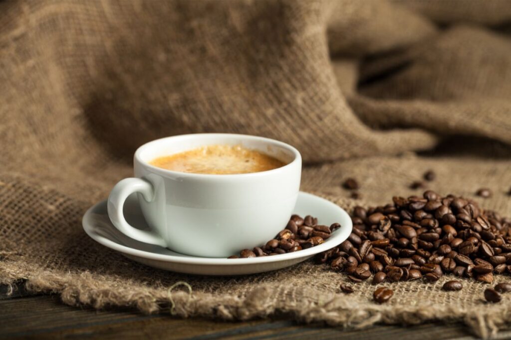 A white cup of coffee with frothy crema sits on a saucer next to a pile of coffee beans, all placed on coarse hessian fabric—perfect for rustic coffee photography.
