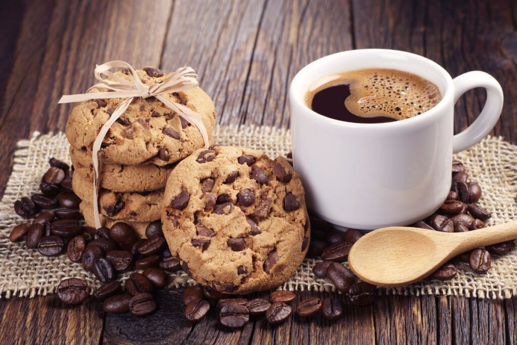 A white mug of coffee sits on a hessian cloth next to chocolate chip biscuits, some tied with string, surrounded by scattered coffee beans and a wooden spoon on a rustic wooden table—perfect for cosy food photography.