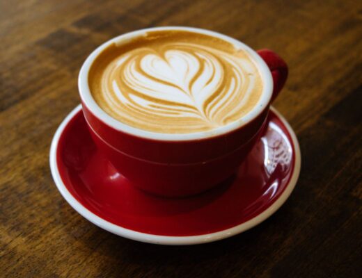 A red cup and saucer filled with a latte, featuring heart-shaped latte art on top, sits on a wooden table—capturing the beauty of coffee through photography.