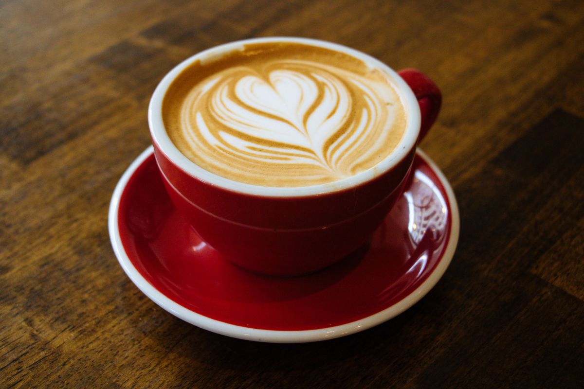 A red cup and saucer filled with a latte, featuring heart-shaped latte art on top, sits on a wooden table—capturing the beauty of coffee through photography.