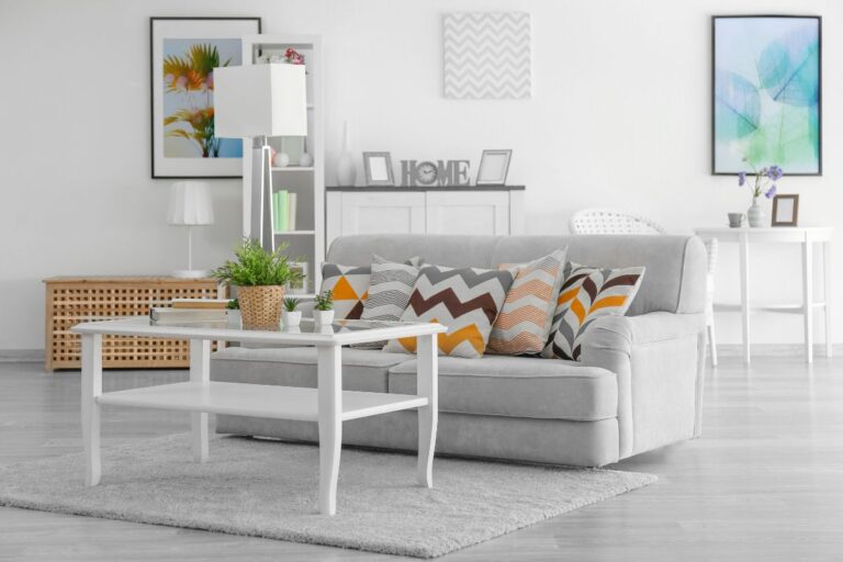Modern living room with a light grey sofa, colourful chevron and geometric cushions, white coffee table with plants, a floor lamp, and wall art décor. This bright and airy space reflects true value home style.