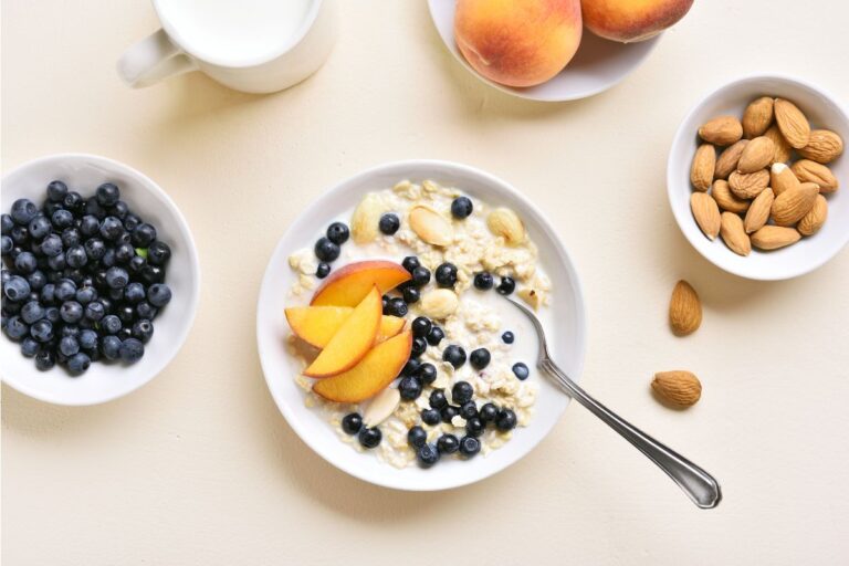 A bowl of porridge oats topped with blueberries, peach slices, and almonds sits on a table, surrounded by bowls of fresh blueberries, almonds, whole peaches, and a mug of milk. A spoon rests in the porridge bowl.