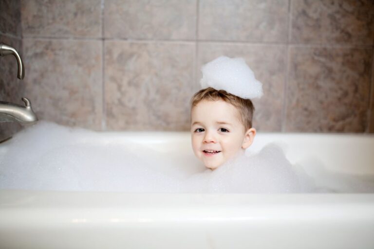 A young child smiles while sitting in a family bathroom bath filled with bubbles, with a playful pile of bubbles on their head. The tiled walls and a silver tap are visible on the left.
