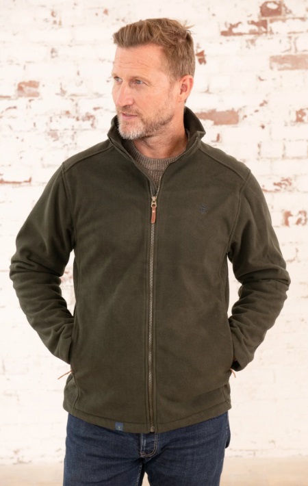 A man with short light brown hair and a trimmed beard stands in front of a white brick wall, wearing a dark green zip-up jacket and blue jeans. He looks to the side as if lost in thought before starting a ramble, hands tucked into his jacket pockets.