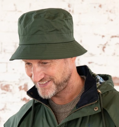 A smiling man with a grey beard wears a dark green bucket hat, ready for a ramble, along with a green jacket with a hood and a brown jumper. The background is a white brick wall with some patches of exposed brick.