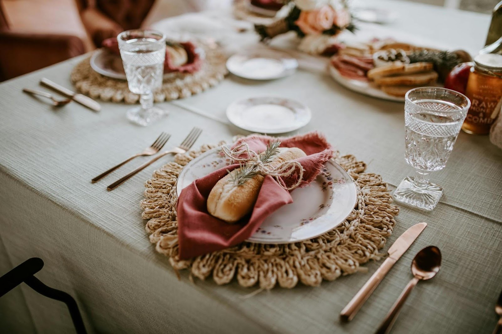 A close-up of an elegant wedding table setting with a plate holding a bread roll wrapped in a red serviette, tied with twine and rosemary. Crystal glasses, gold-coloured cutlery, and decorative place mats adorn the table.