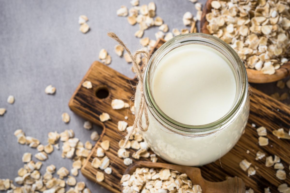A jar of creamy oat milk sits on a wooden board, surrounded by scattered porridge oats and a wooden bowl filled with oats, on a grey surface.