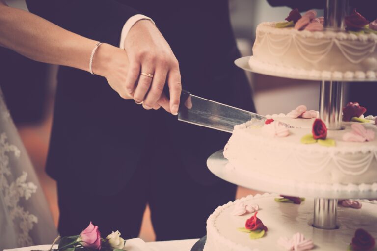 A bride and groom, both wearing wedding rings, cut a tiered white wedding cake decorated with pink and red sugar roses at their wedding. The bride’s lace sleeve and the groom’s suit are visible as they share this special moment.