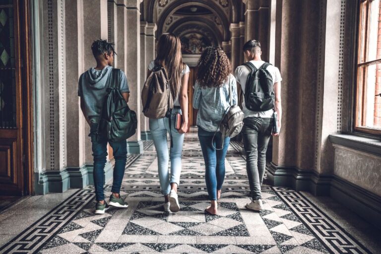 Four students with rucksacks walk down a corridor with ornate columns and patterned tiled floor, seen from behind. The setting appears academic, suggesting they are exploring a historic university campus.