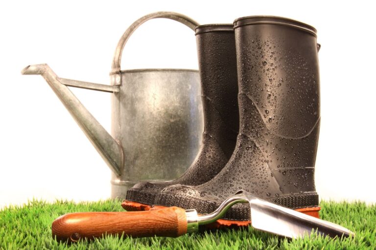 A pair of wet black rubber wellies, a metal watering can, and a hand trowel with a wooden handle sit on bright green garden grass against a white background.