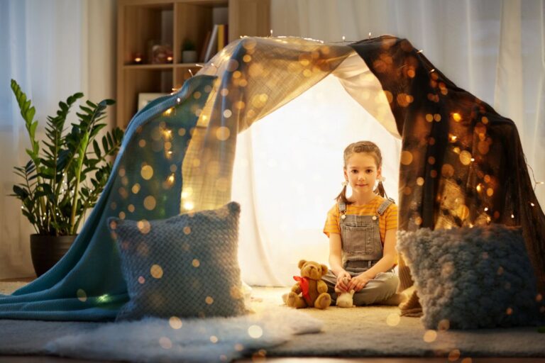 A young girl enjoys rainy day activities inside a cosy homemade blanket fort decorated with fairy lights, holding a teddy bear. Soft cushions are scattered on the floor, warm light glows around her, and houseplants and shelves fill the background.