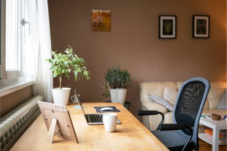 A home office designed for working from home features a wooden desk with a laptop, mouse mat, coffee mug, potted plant, and photo frame by the window. An office chair, sofa, and wall art complete the comfortable workspace.
