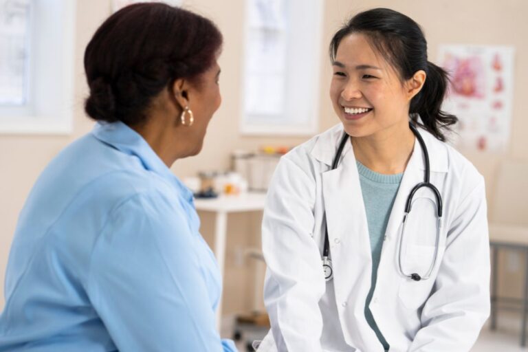 A smiling female doctor wearing a white coat and stethoscope talks with a patient in a blue shirt during routine health check-ups in a bright medical surgery.