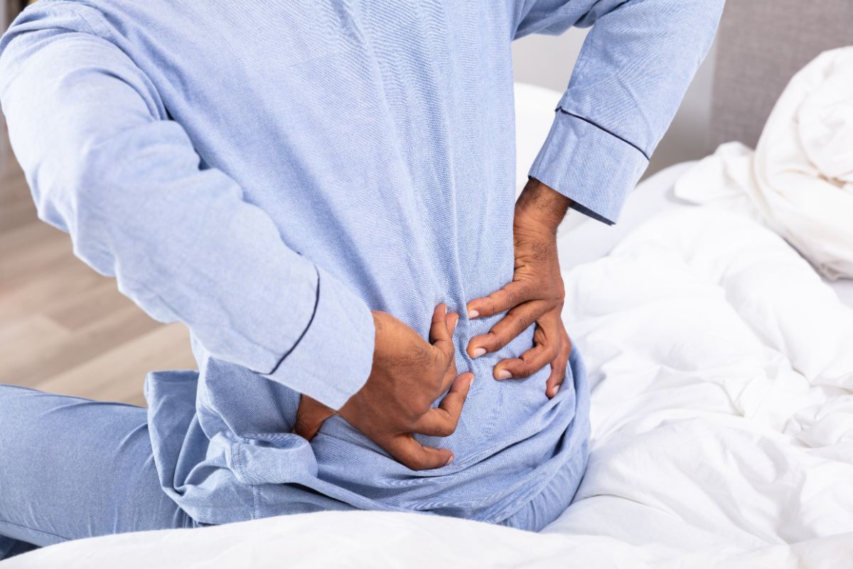 A person in blue pyjamas sits on a bed, facing away, holding their lower back with both hands as if experiencing discomfort—possibly due to an unsupportive mattress.