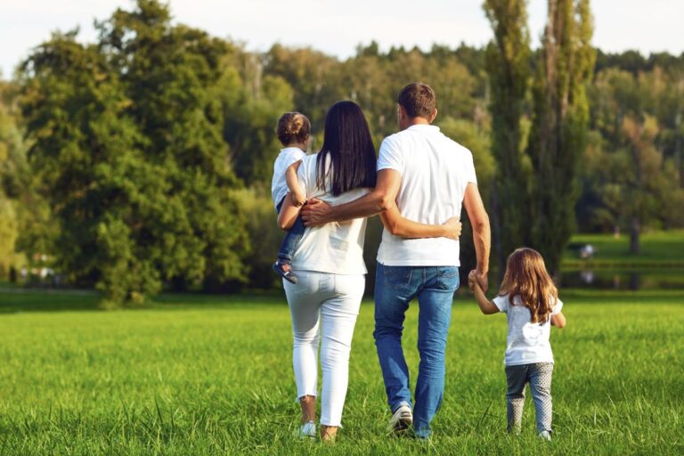 A family of four, seen from behind, walks hand in hand through a grassy field. The parents hold their children, enjoying outdoor activities on a sunny day surrounded by green trees and nature.