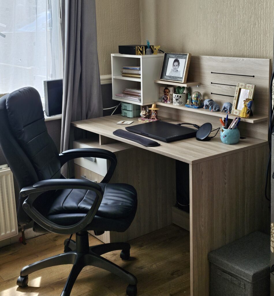 A tidy work-from-home office workspace with a black leather chair, wooden desk, monitor, keyboard, mouse, stationery, and shelves holding framed photos, toys, and small decorations near a window with grey curtains.