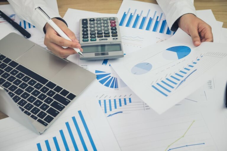 A person works at a desk with a laptop, calculator, pen, and various financial charts and graphs, analysing data and making notes on printed reports as part of effective financial management.