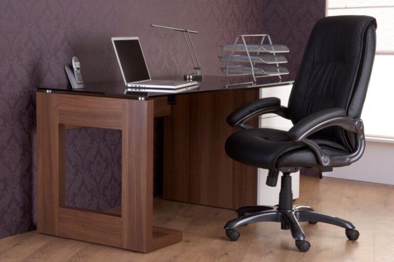 A modern work from home office setup features a dark wooden desk, black leather chair, laptop, desk lamp, phone, metal file organiser, and stylish patterned purple wallpaper in the background.