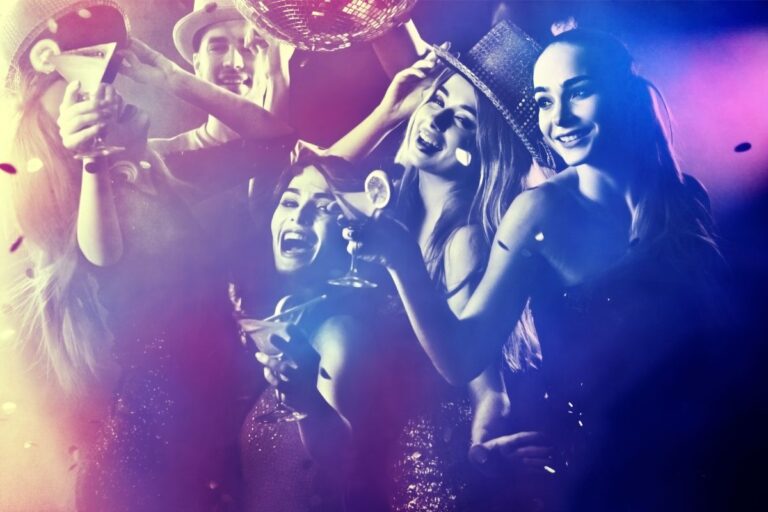 A group of young adults dressed up and smiling, holding cocktails and dancing under a disco ball at a lively cocktail party, with colourful lights and confetti in the air.
