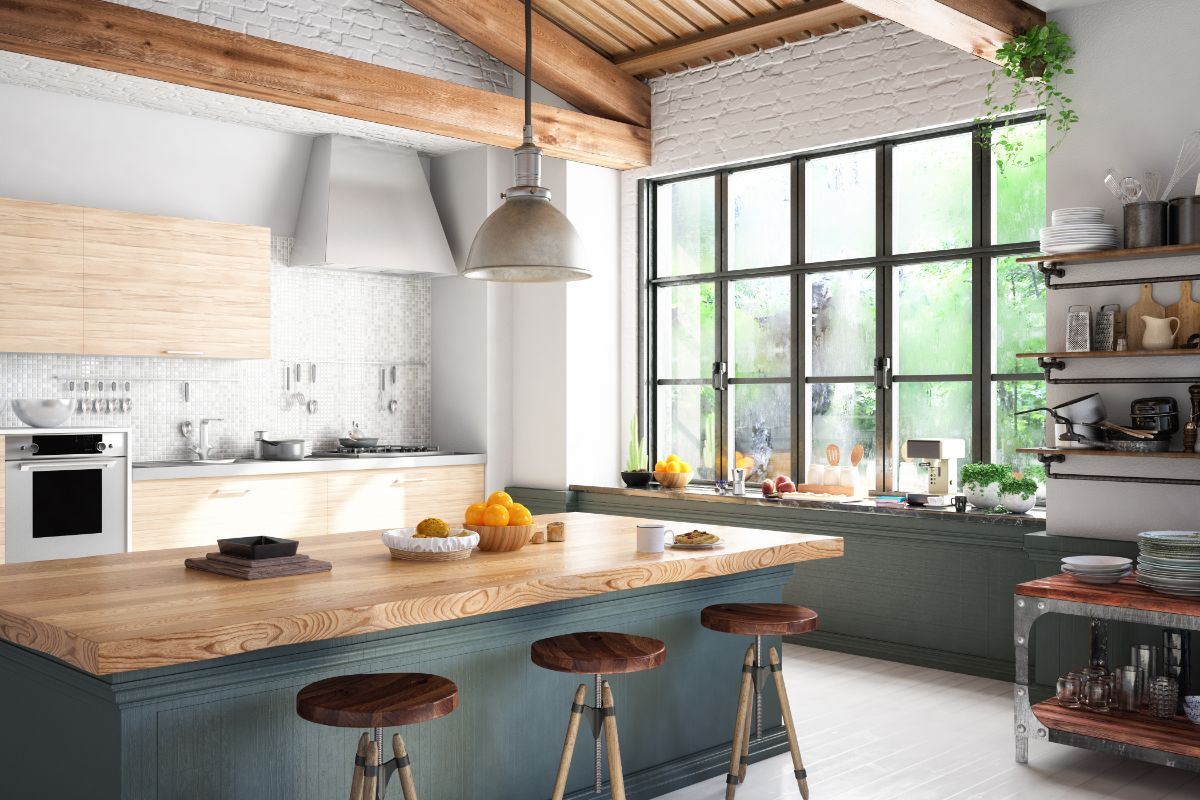 Bright, modern kitchen with a large wooden island, three stools, open shelving, and a big window letting in natural light. Fruit and kitchen items sit on the worktop and shelves. Exposed beams and brick add rustic charm to this modern kitchen.