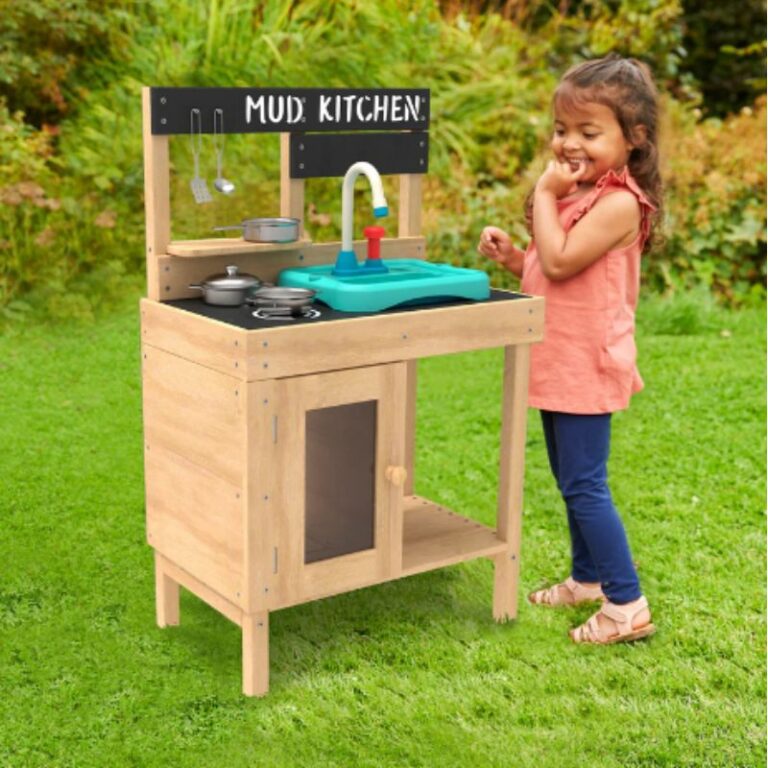 A young girl stands next to a wooden play kitchen labelled "Mud Kitchen" in a grassy outdoor area, embracing nature as she tastes or smells something with a playful expression. Pots, pans, and utensils are arranged on the play kitchen.