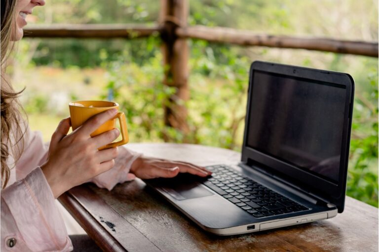 A person sitting at a wooden table outdoors holds a yellow mug in one hand and uses the other hand to type on a laptop, enjoying remote working with greenery visible in the background.