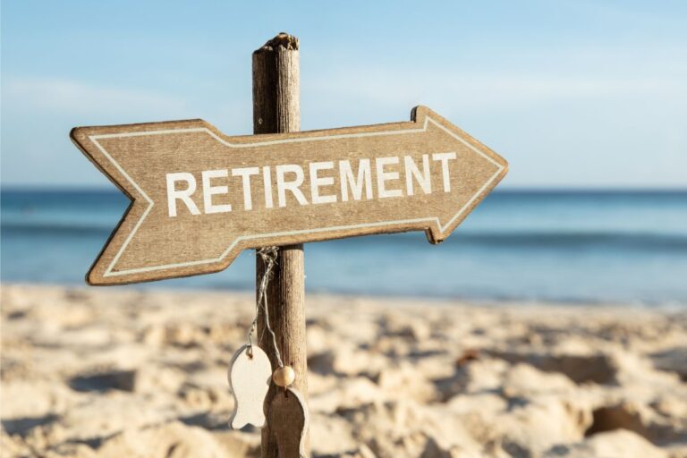 A wooden arrow sign reading "RETIREMENT" stands on a sandy beach, with the sea and blue sky in the background—a perfect symbol for a peaceful retirement getaway. Small decorative tags hang from the signpost.
