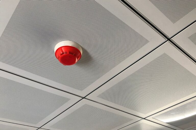 Red and white smoke detector, one of the essential safety devices, mounted on a perforated, square-patterned ceiling.