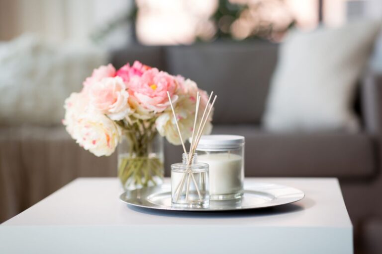 A small glass jar with reed diffusers releases a gentle aroma beside a white candle and a vase of pink and white flowers on a tray atop a white table, with a blurred sofa and cushions in the background.