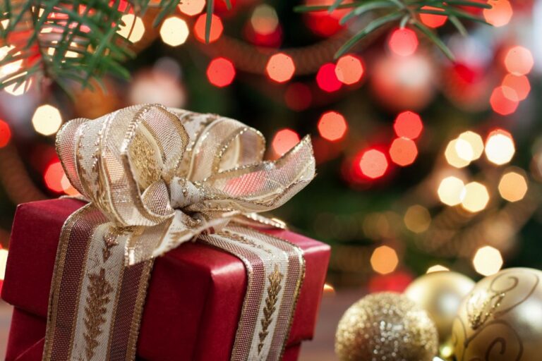 A red gift box with a gold ribbon and bow sits in front of a blurred background of red, gold, and white holiday lights—perfect for inspiring Christmas gift ideas—whilst pine branches and gold baubles add festive charm nearby.