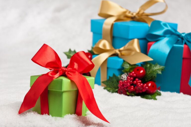 A green gift box with a red ribbon sits on snow in front of blue gift boxes with gold and blue ribbons, evoking a meaningful Christmas. A small holiday decoration with red berries and green leaves completes the festive scene.