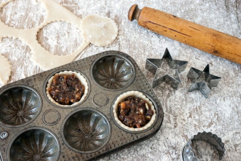 A vintage muffin tray with partly filled pastry cases sits on a floured surface, surrounded by rolled pastry, star-shaped biscuit cutters, a round cutter, and a wooden rolling pin—perfect for festive Christmas baking.