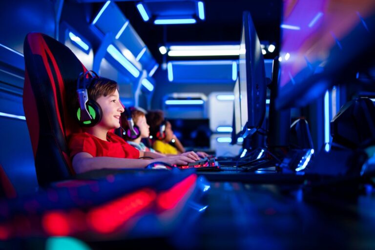Three children wearing headsets sit in gaming chairs, smiling and playing video games on computers in a room lit with blue and neon lights, creating a futuristic gaming atmosphere.